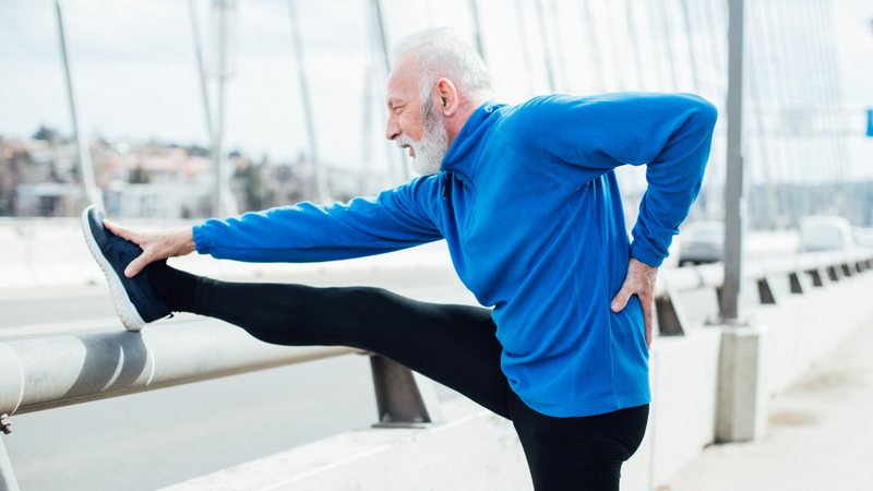 Homem idoso usando roupas esportivas com a mão nas costas enquanto se alonga - RgStudio/Getty Images